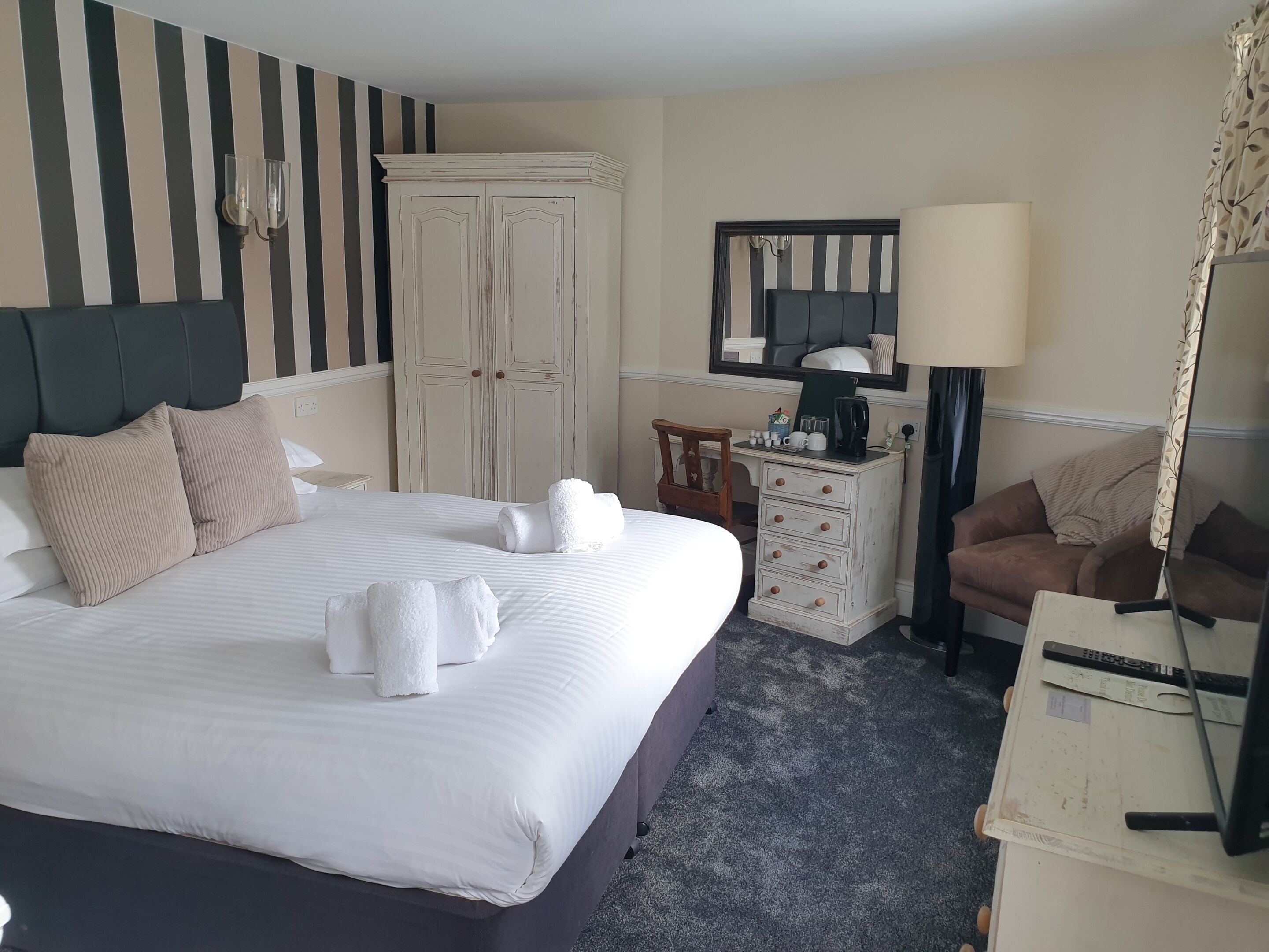 A hotel room with a large bed, a wardrobe, a small desk with a lamp, a chair, and a sofa.