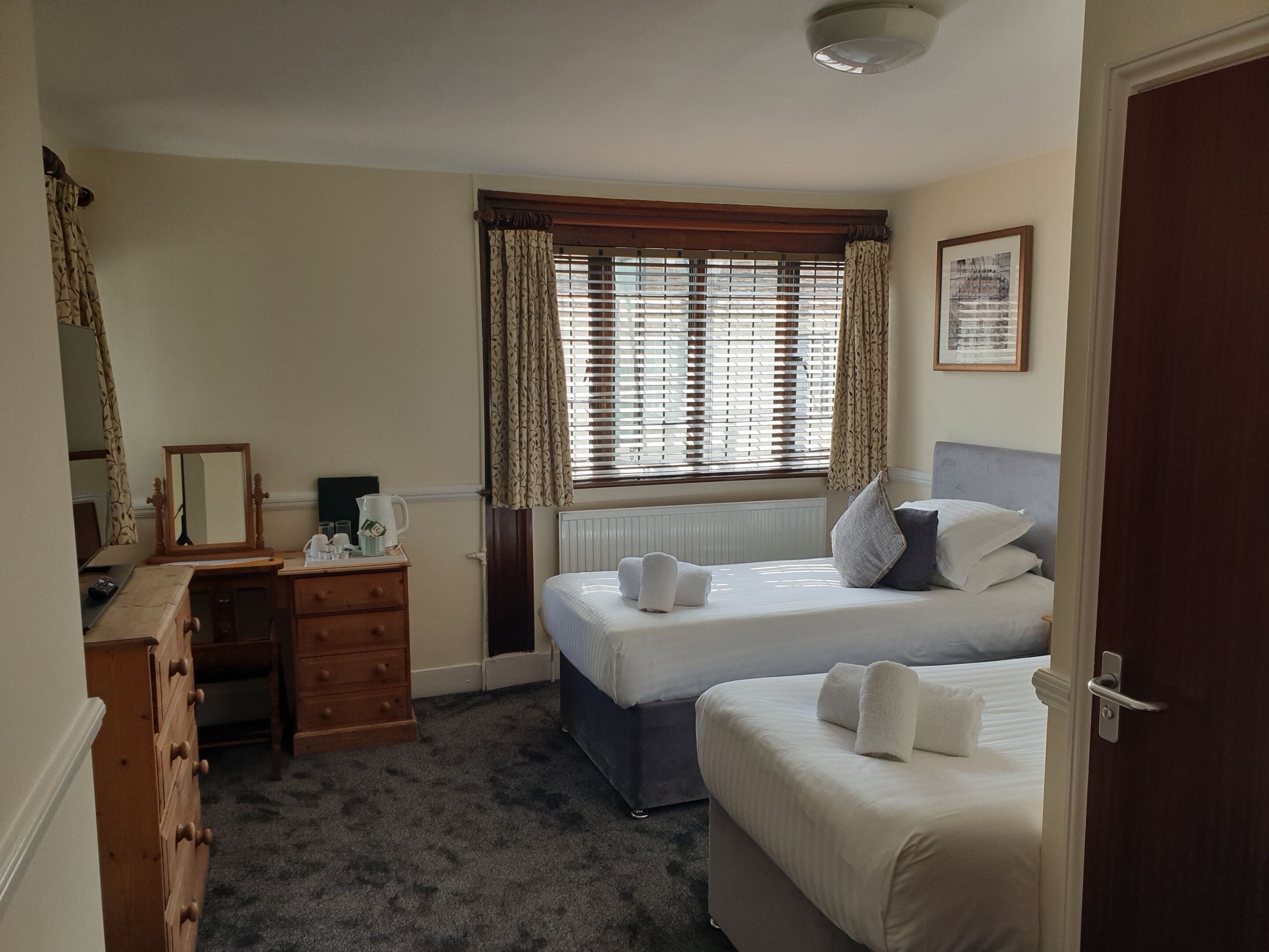 A bedroom with two beds, a dresser, a desk, a radiator, and a window with curtains.