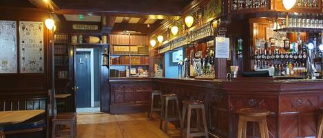 A bar with wooden flooring, bar stools, and a variety of bottles and glasses on shelves.