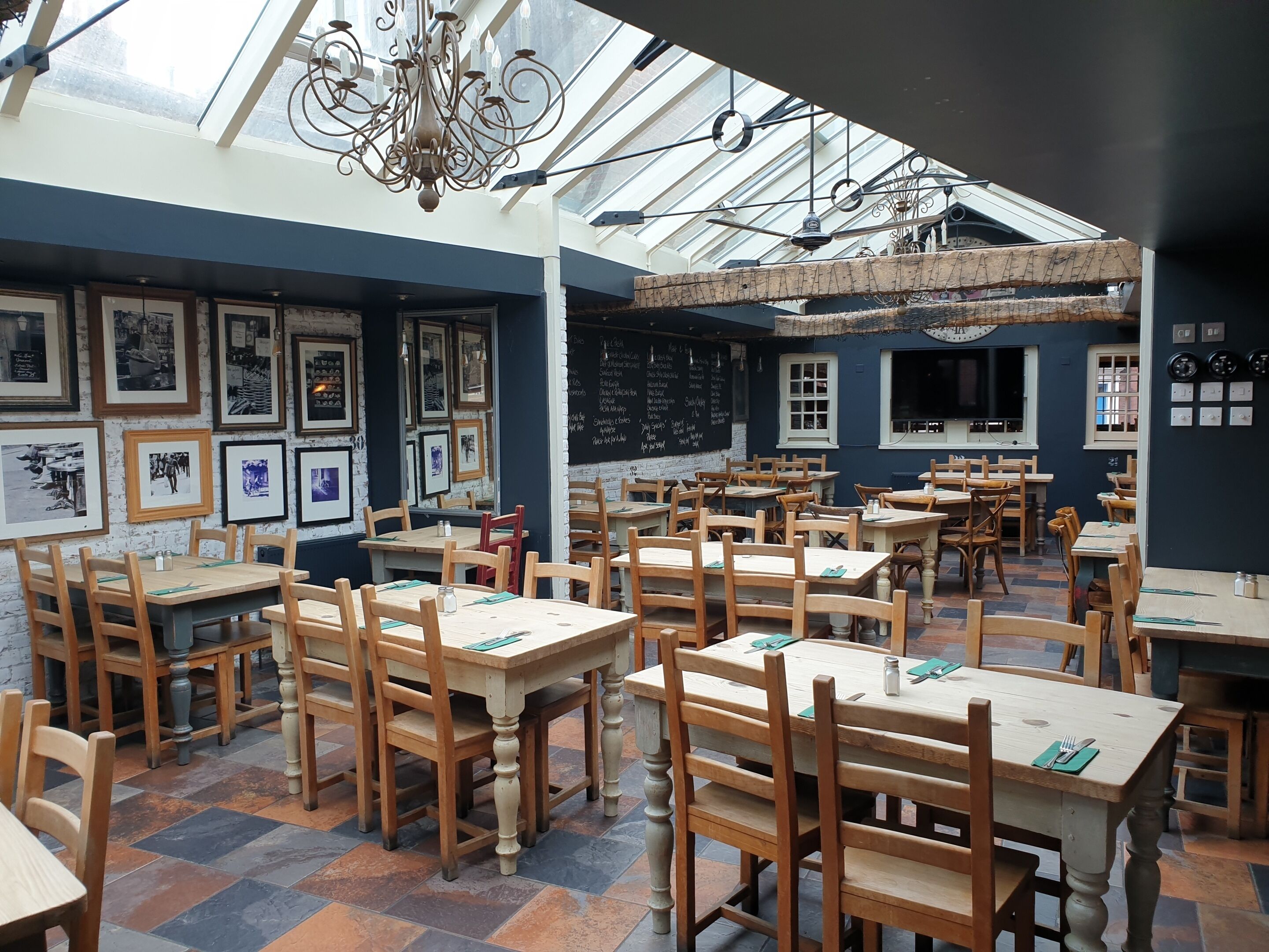 Een restaurant met houten tafels en stoelen, lijstjes met foto's aan de muren en een menukaart op een schoolbord.