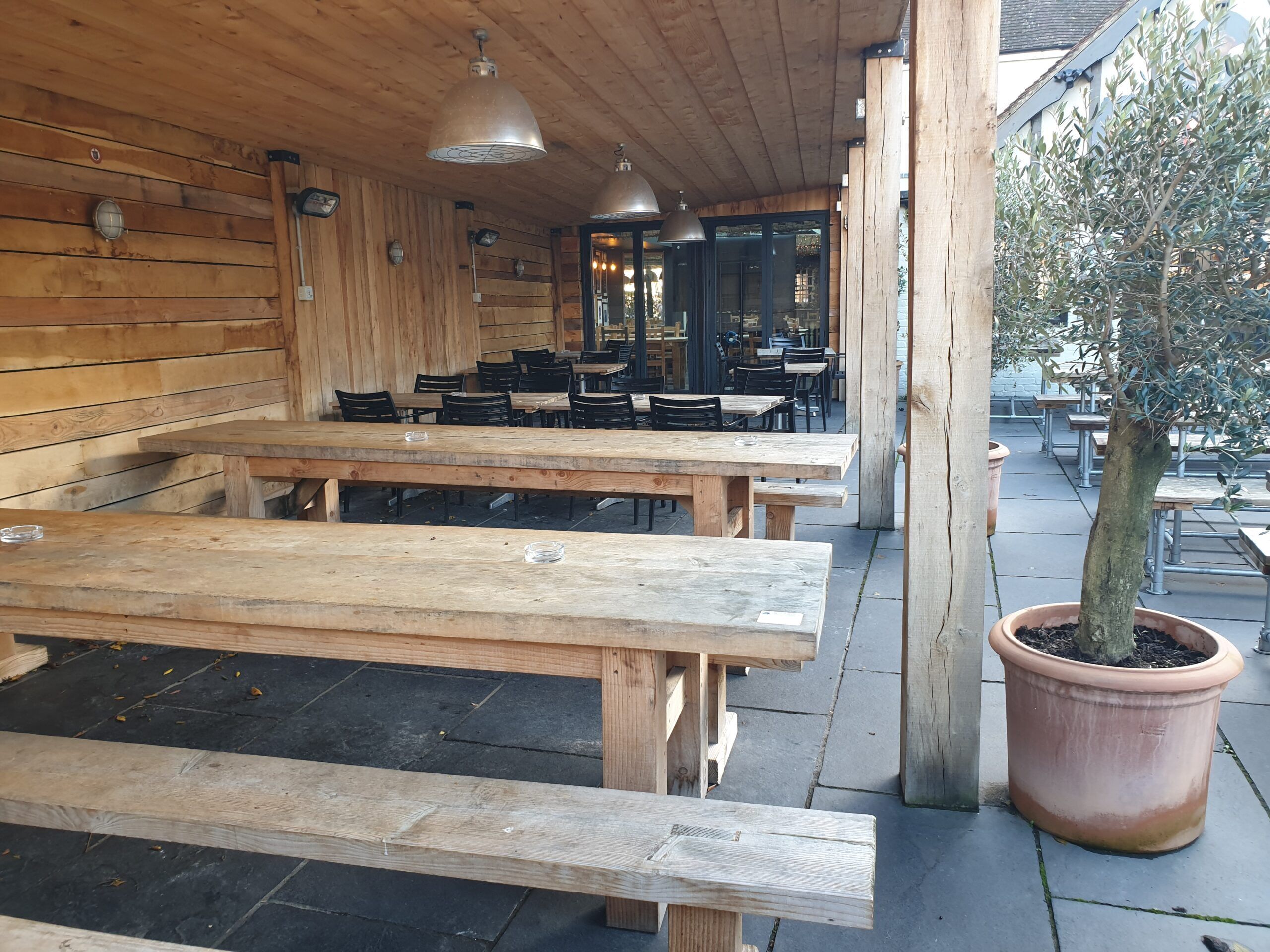 An outdoor seating area with wooden benches and tables, a potted tree, and a building with wooden walls and hanging lights.