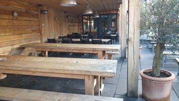 An outdoor seating area with wooden benches and tables, a potted tree, and a building with wooden walls and hanging lights.