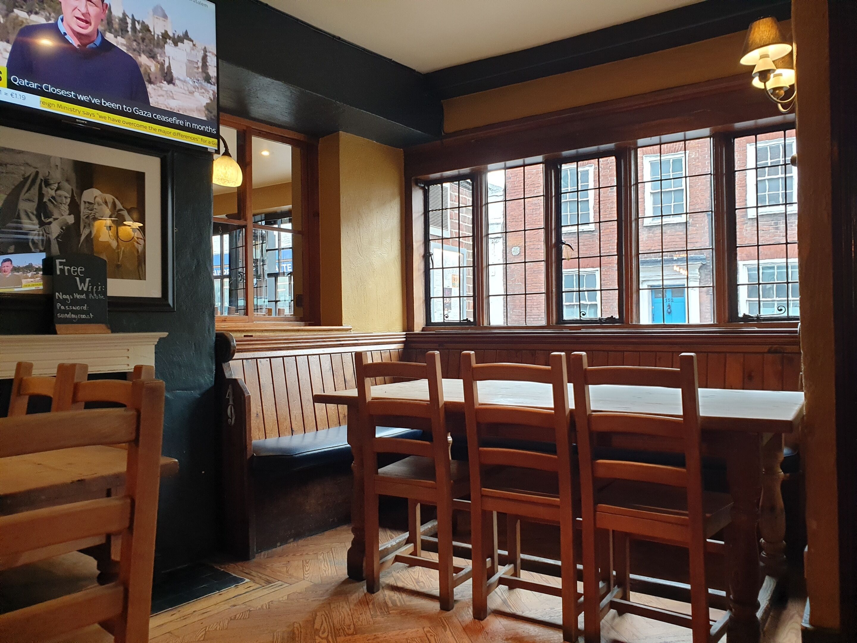 L’intérieur d’un restaurant avec des tables et des chaises en bois, une grande fenêtre donnant sur des immeubles et un téléviseur diffusant les actualités.