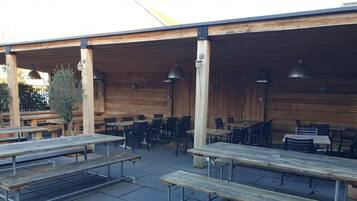 Un patio al aire libre con mesas y bancos de madera, barandillas metálicas y luces colgantes.