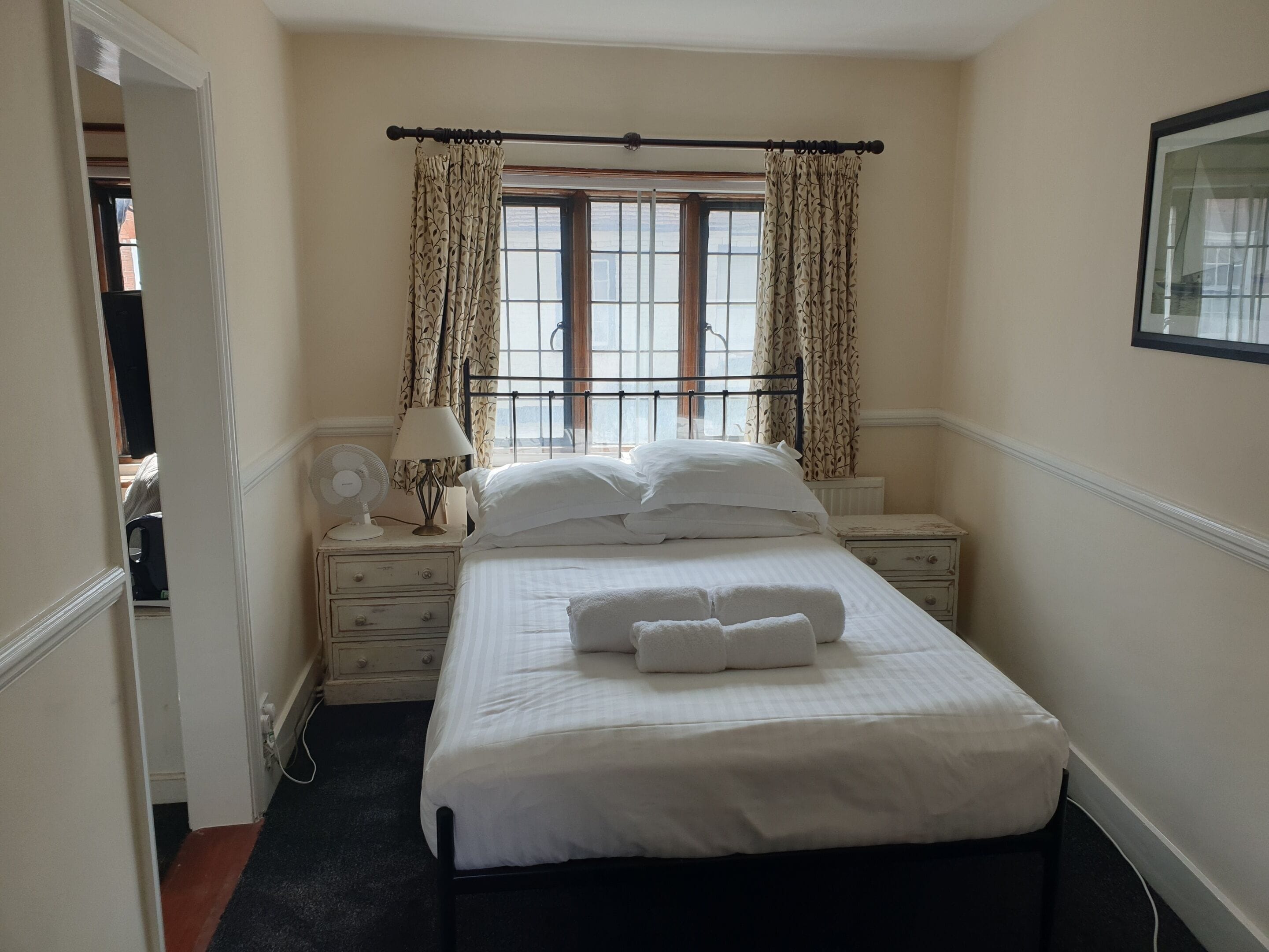 A bedroom with a bed, a nightstand with a lamp, a window with curtains, and a radiator.