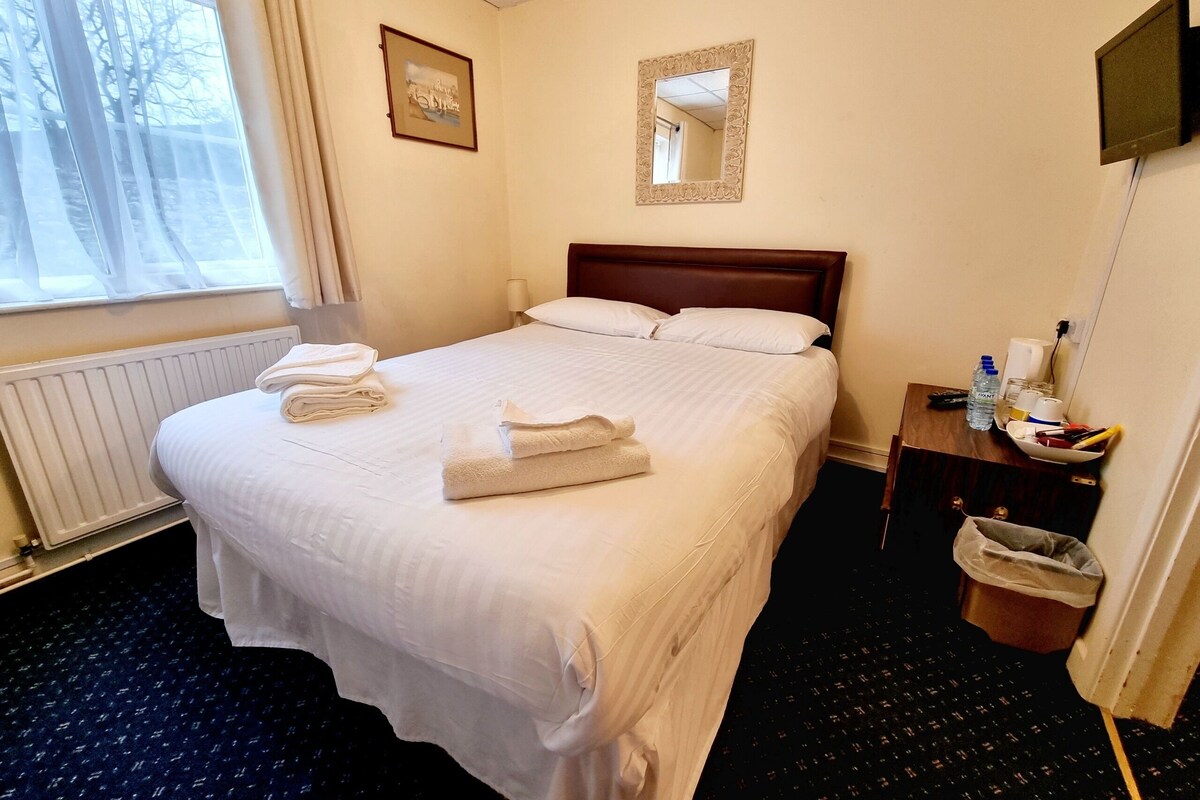 Une chambre d’hôtel avec un lit bien fait, une table de chevet avec une lampe, un téléviseur fixé au mur et une fenêtre avec des rideaux.