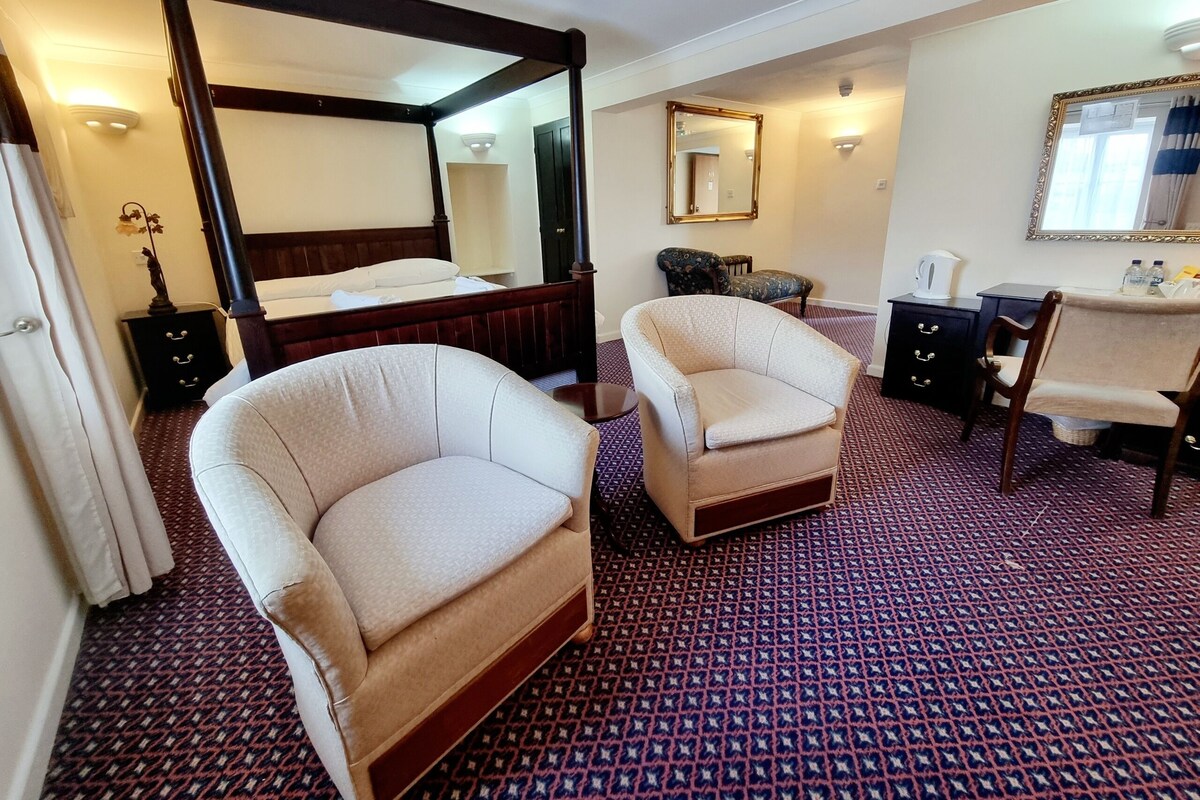 Une chambre d’hôtel avec un lit à baldaquin, deux fauteuils, un bureau avec une chaise, un miroir et une moquette à motifs.