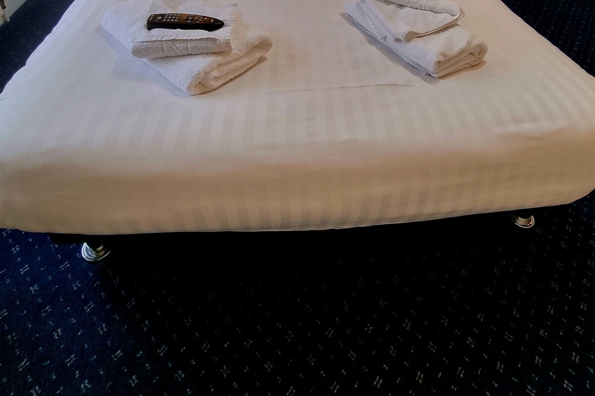 Un lit bien fait, avec des draps blancs, deux serviettes pliées et une télécommande posée sur le côté gauche.