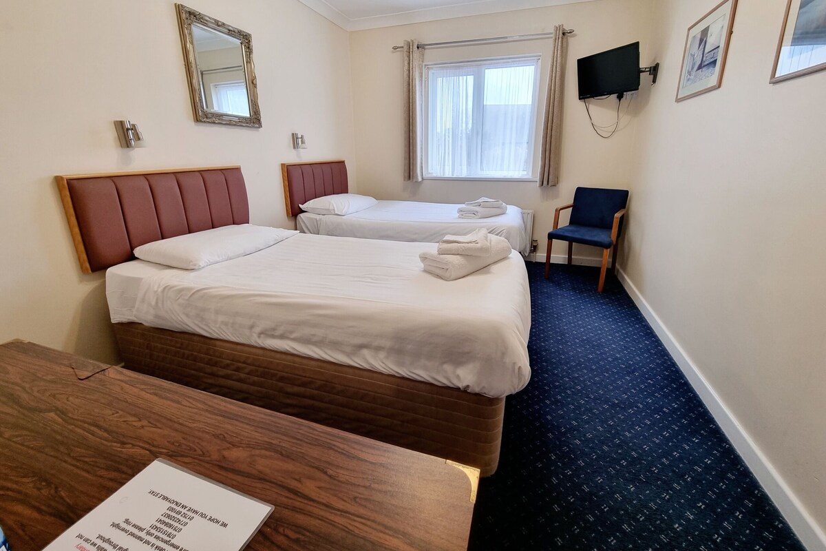 Une chambre d’hôtel avec deux lits, un bureau en bois, une chaise, une télévision et une fenêtre avec des rideaux.