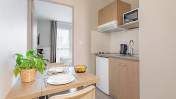 Een praktische keuken met een magnetron, waterkoker en een kleine koelkast. Er is een eettafel met stoelen, een plant en een schaal met fruit.
