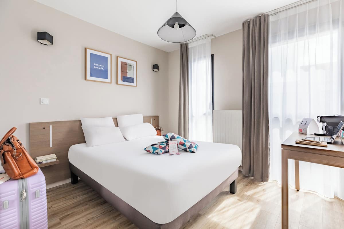 Une chambre d’hôtel avec un grand lit, un bureau équipé d’un ordinateur, une lampe et une fenêtre avec des rideaux.