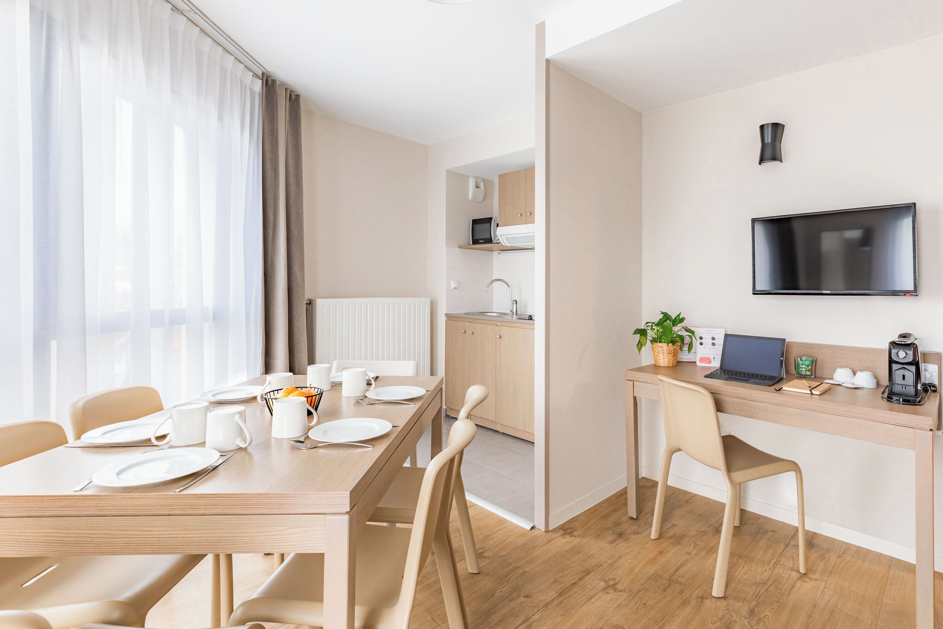 A modern hotel room with a dining area, kitchenette, and a TV mounted on the wall.