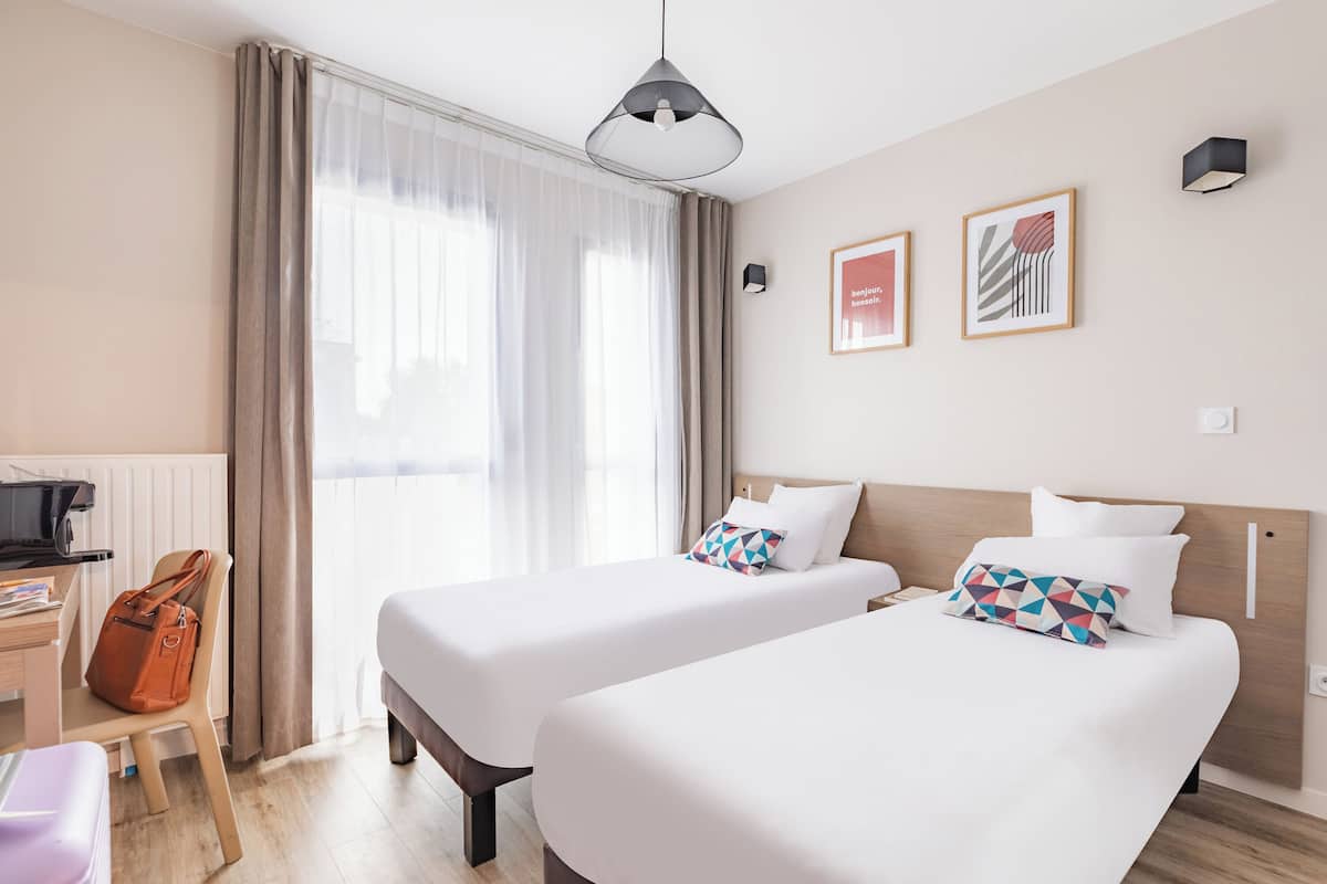 Une chambre d’hôtel avec deux lits, un bureau, une chaise et une fenêtre avec des rideaux.
