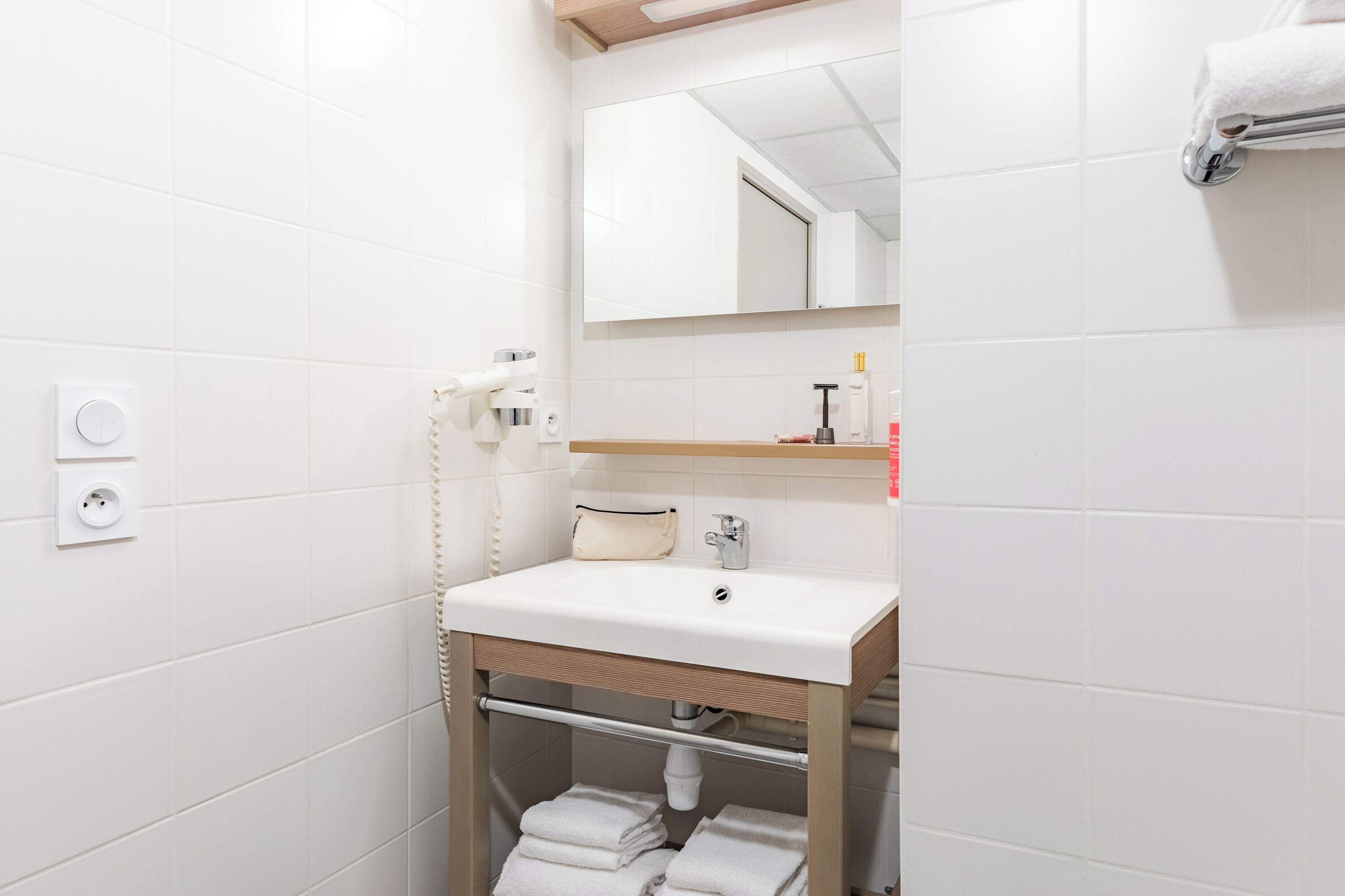 A modern bathroom with white tiles, a sink with a mirror above, a towel rack with towels, and a small shelf with toiletries.