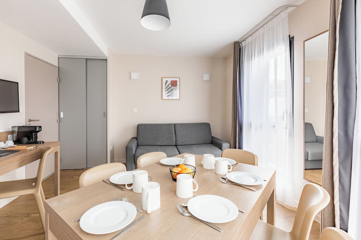 Un espace repas moderne avec une table en bois dressée pour quatre personnes, un canapé gris en arrière-plan et un coin cuisine sur la gauche.