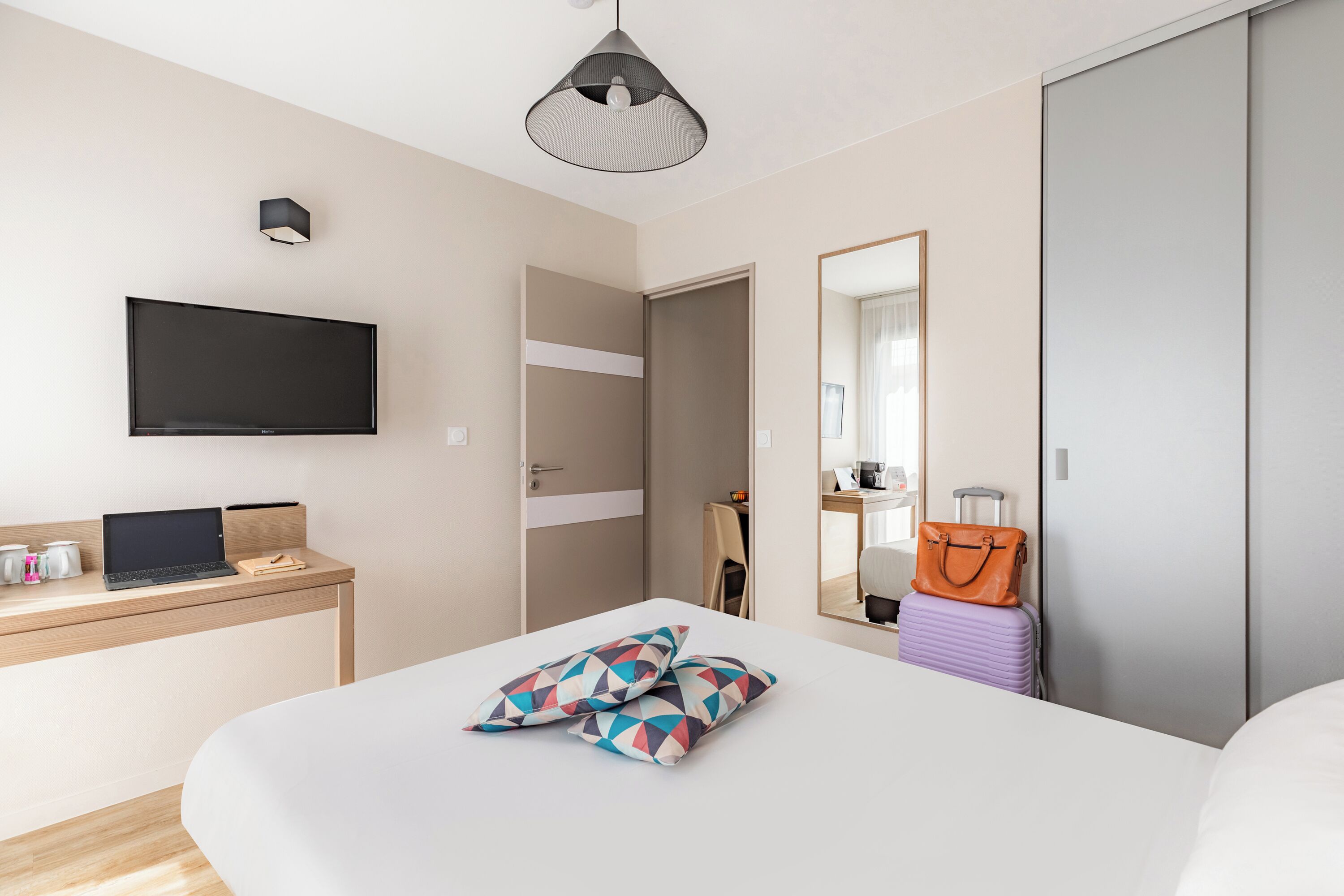Una habitación de hotel moderna con una cama, un escritorio con una computadora portátil, un televisor de pantalla plana, un espejo y una maleta.