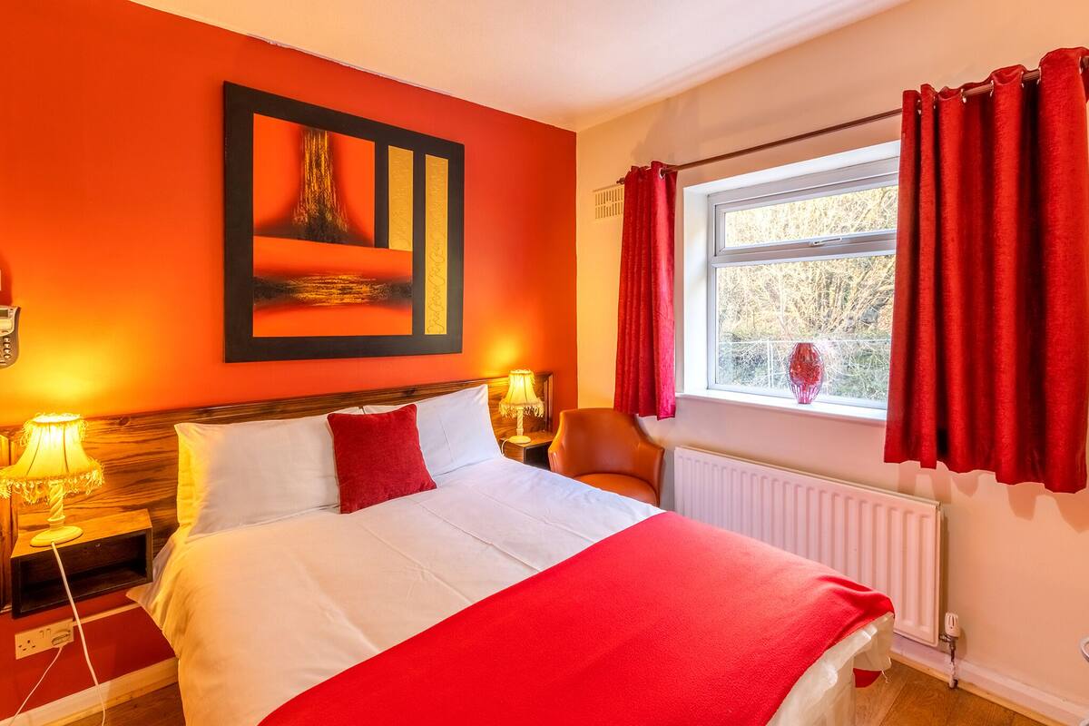 A hotel room with a large bed, red curtains, a window with a view, and a painting on the wall.