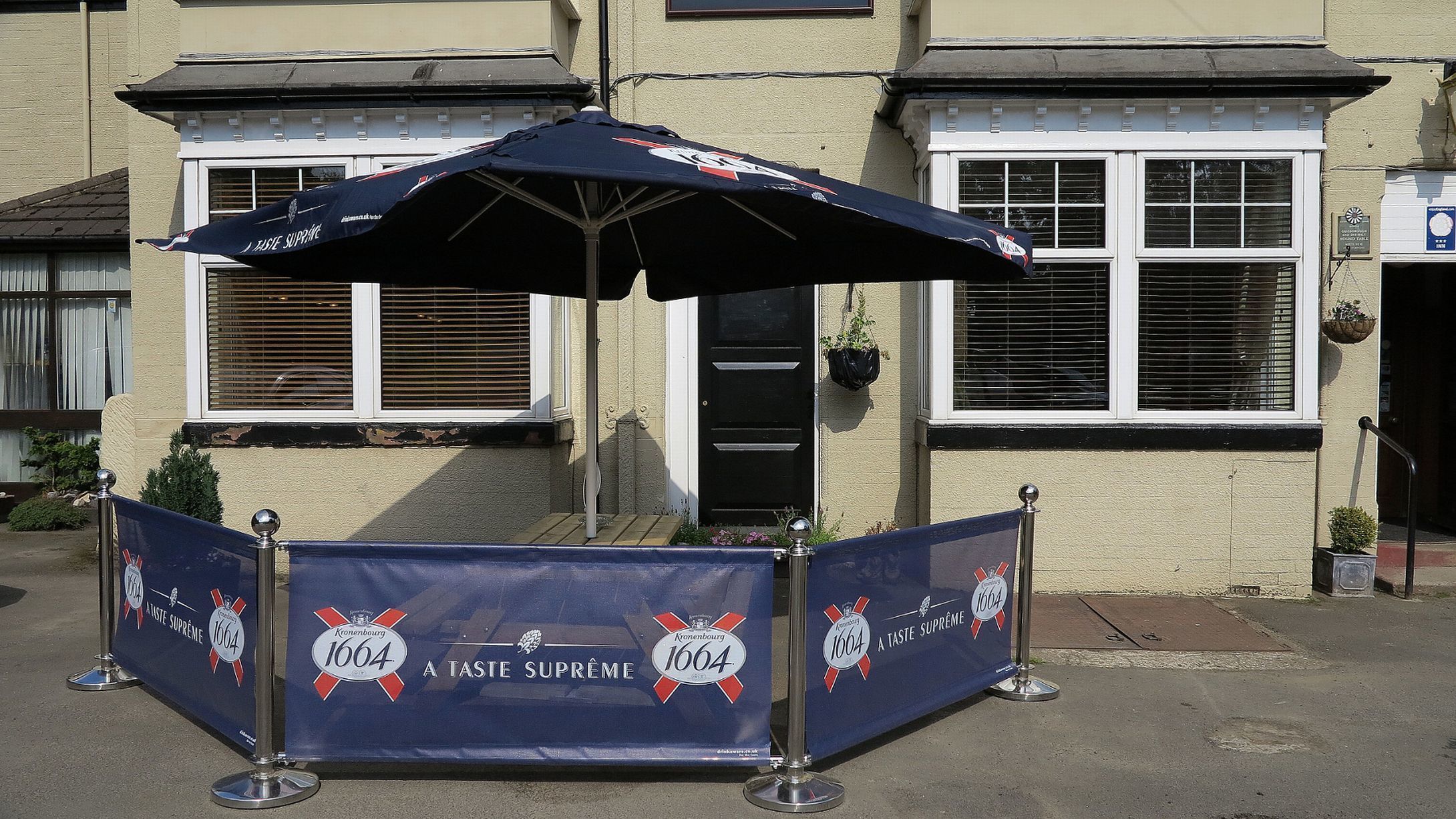 Een blauwe banner met de logo's 'A Taste Supreme' en '1664', een zwarte paraplu en metalen barrières voor een gebouw met ramen en een deur.