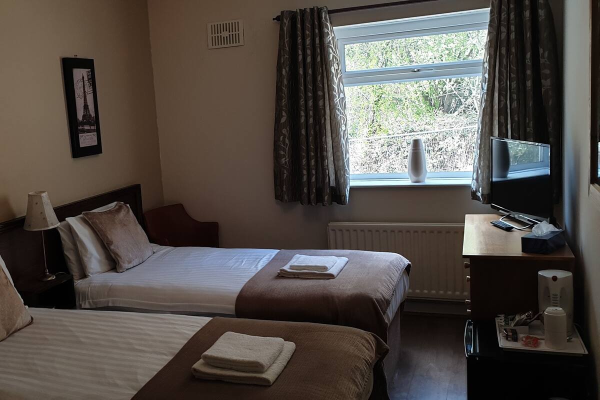 A hotel room with two beds, a desk, a window with curtains, and framed pictures on the wall.