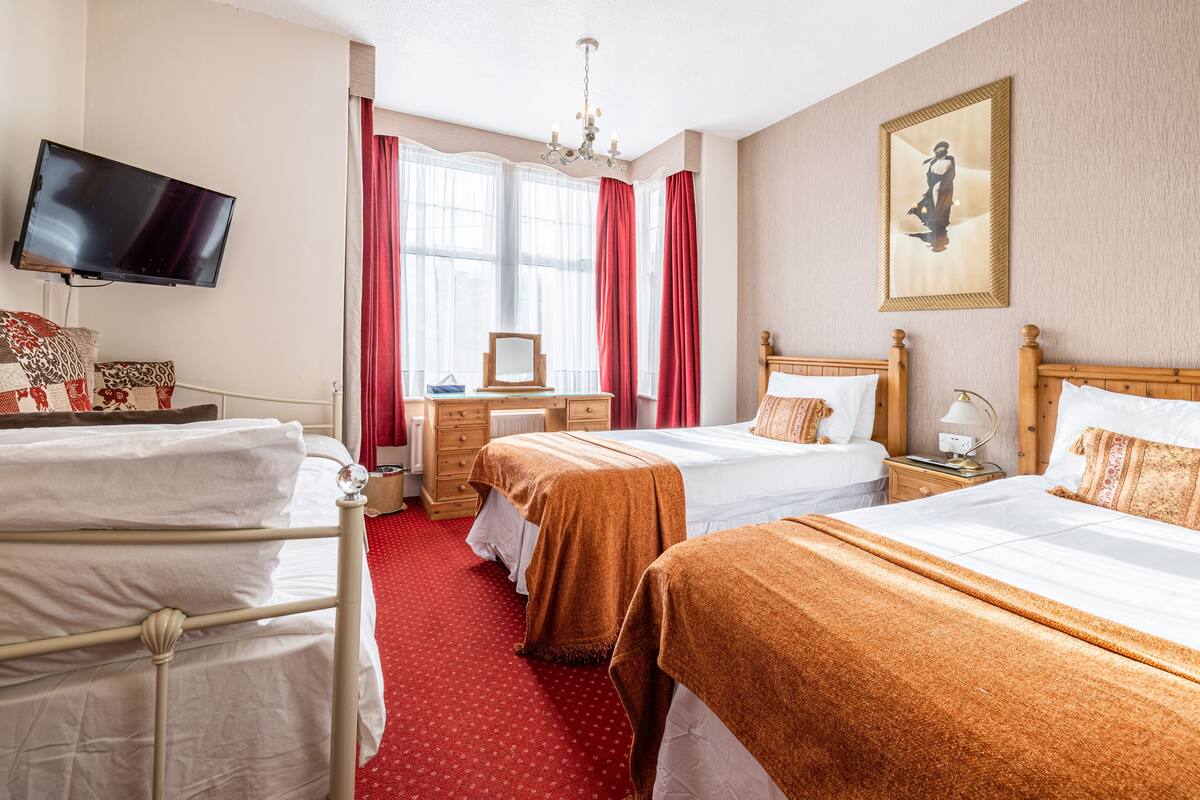 A hotel room with two beds, a TV, a desk, and a large window with red curtains.