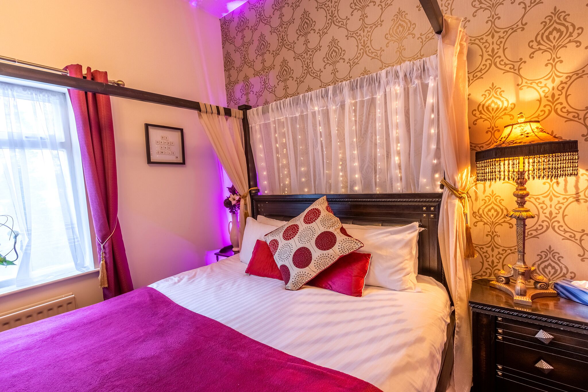 Een netjes opgemaakt bed met een wit en rood gebonden kussen, een paarse plaid en een nachtkastje met een lamp.