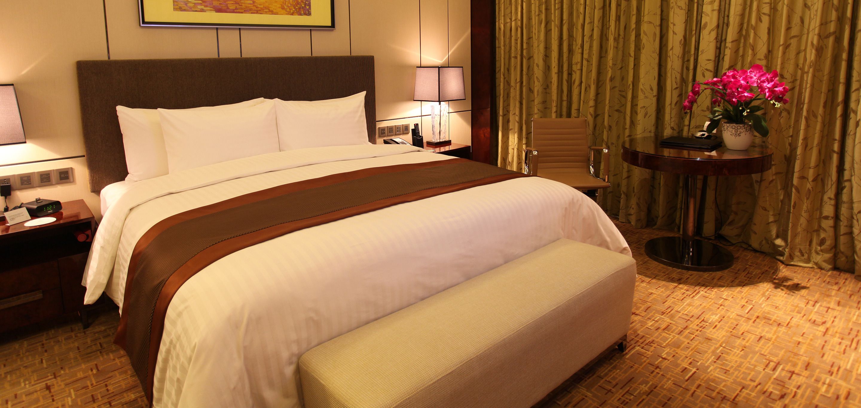 Une chambre d’hôtel avec un grand lit, des tables de chevet, une chaise, une petite table ornée d’un vase de fleurs et un rideau.