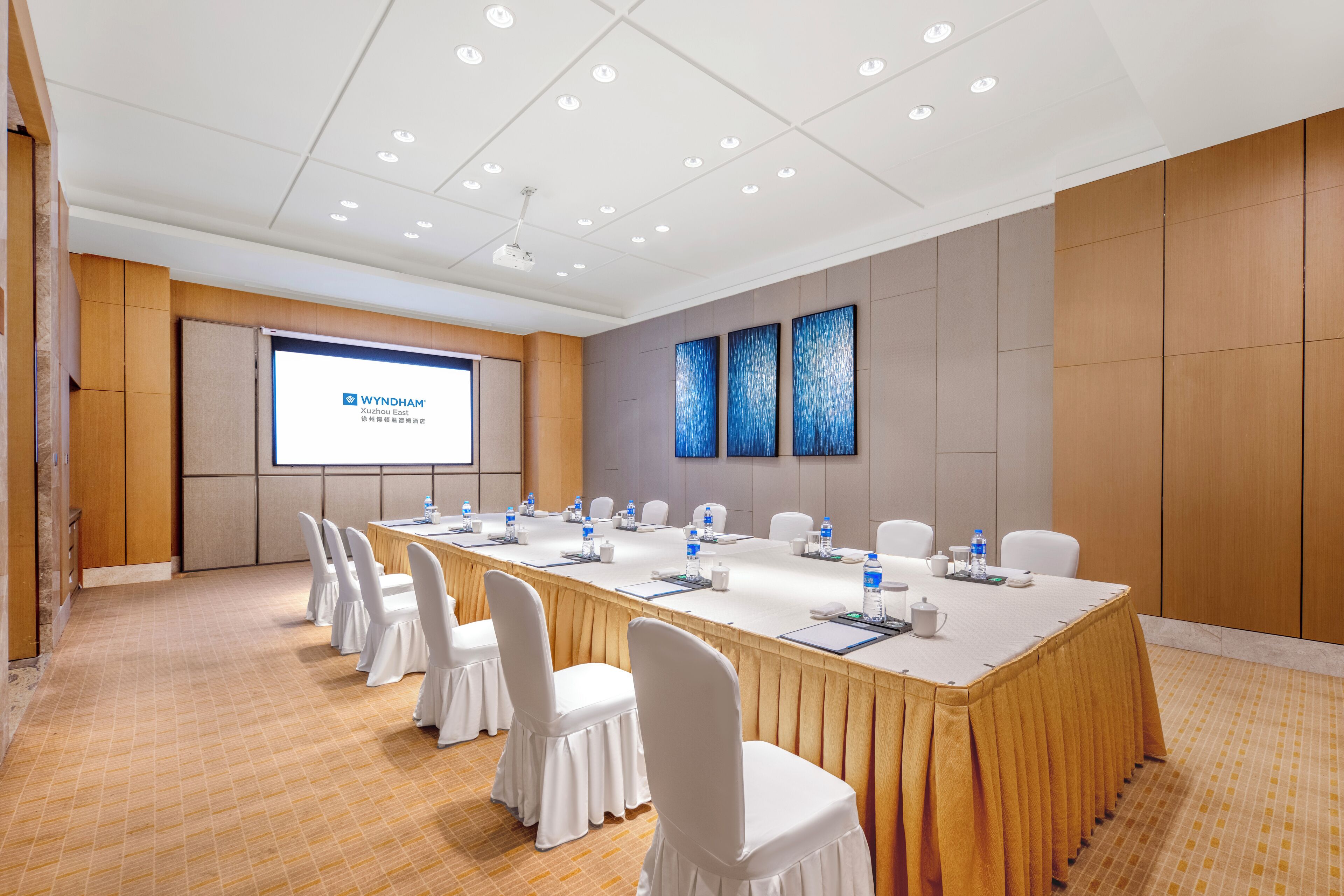 Un salón de conferencias con una larga mesa, sillas blancas y una pantalla mostrando 'WYNDHAM Xuhui East'.