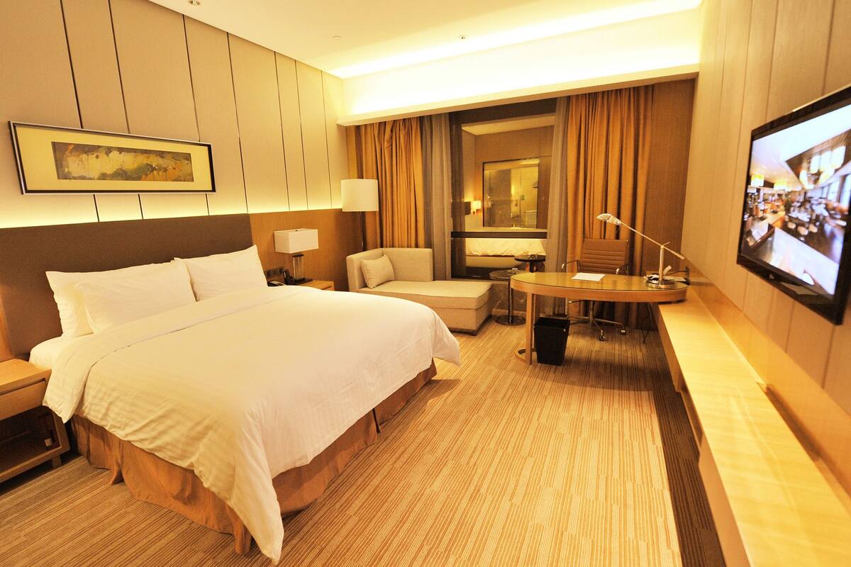 Ein Hotelzimmer mit einem großen Bett, einem Schreibtisch mit Stuhl, einem Fernseher und einem Fenster mit Vorhängen.