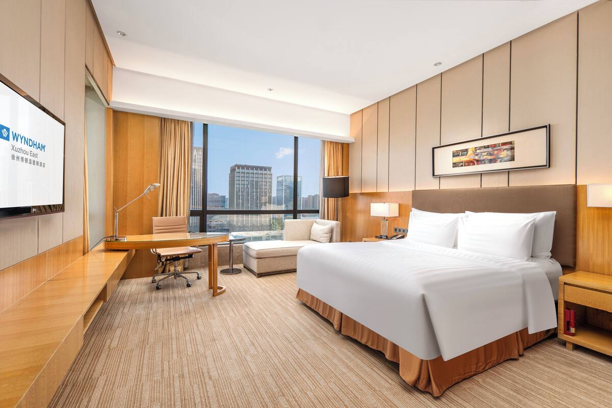 Ein Hotelzimmer mit einem großen Bett, einem Schreibtisch, einem Sessel, einer Couch, einem Fernseher und Blick auf die Stadt.