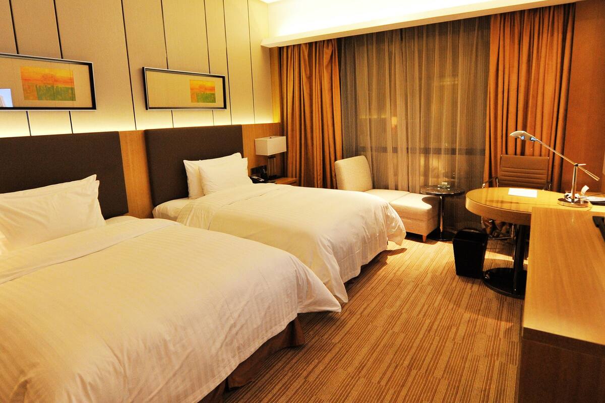 Ein Hotelzimmer mit zwei Betten, einem Schreibtisch mit Lampe, einem Fernseher und einem kleinen Tisch mit Mülleimer.