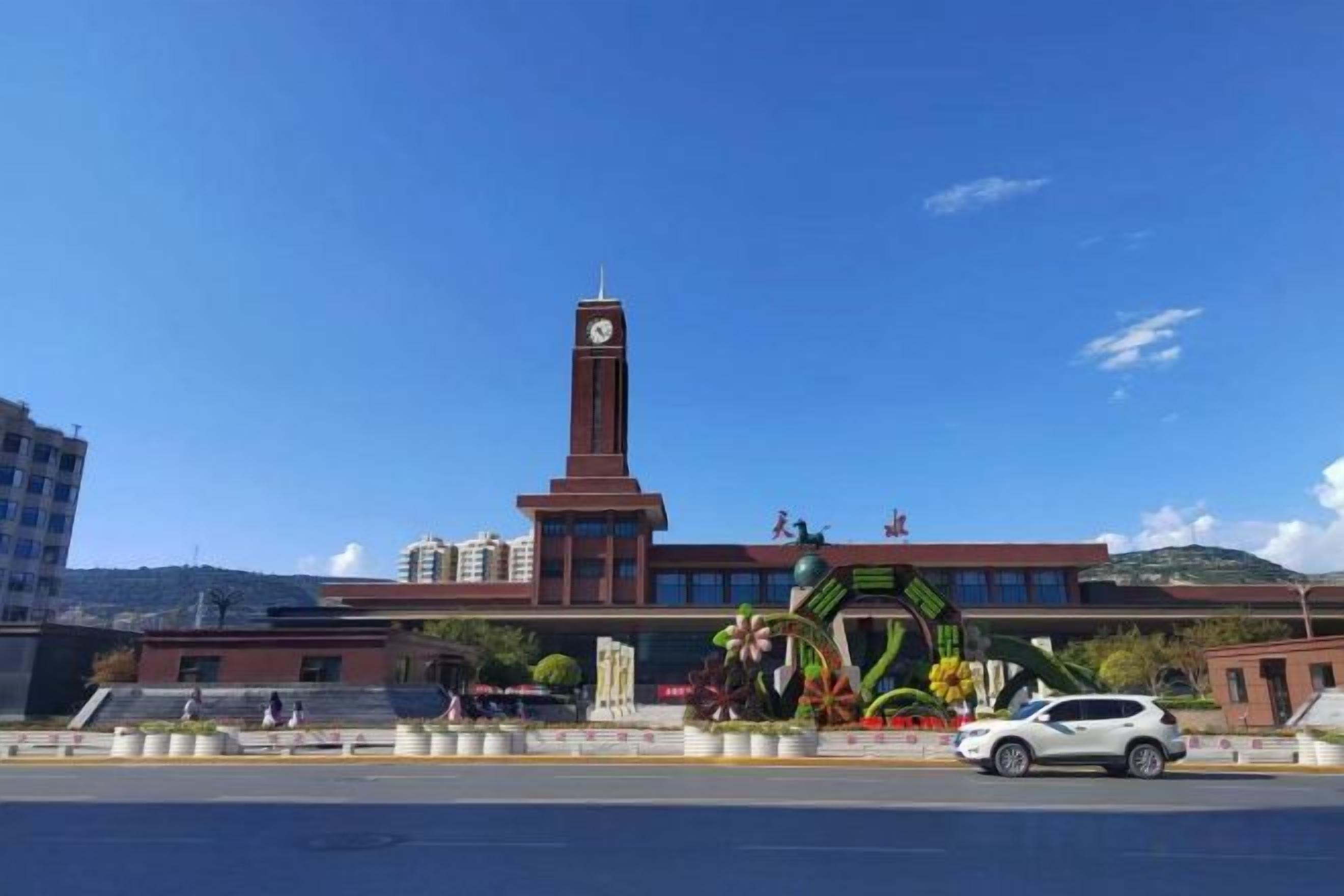 Un edificio con una torre del reloj, una plaza central con una escultura decorativa y un coche estacionado en frente.