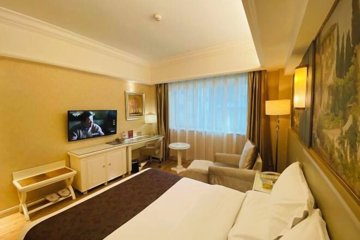 A hotel room with a bed, a desk with a television, a chair, a small table, and a painting on the wall.