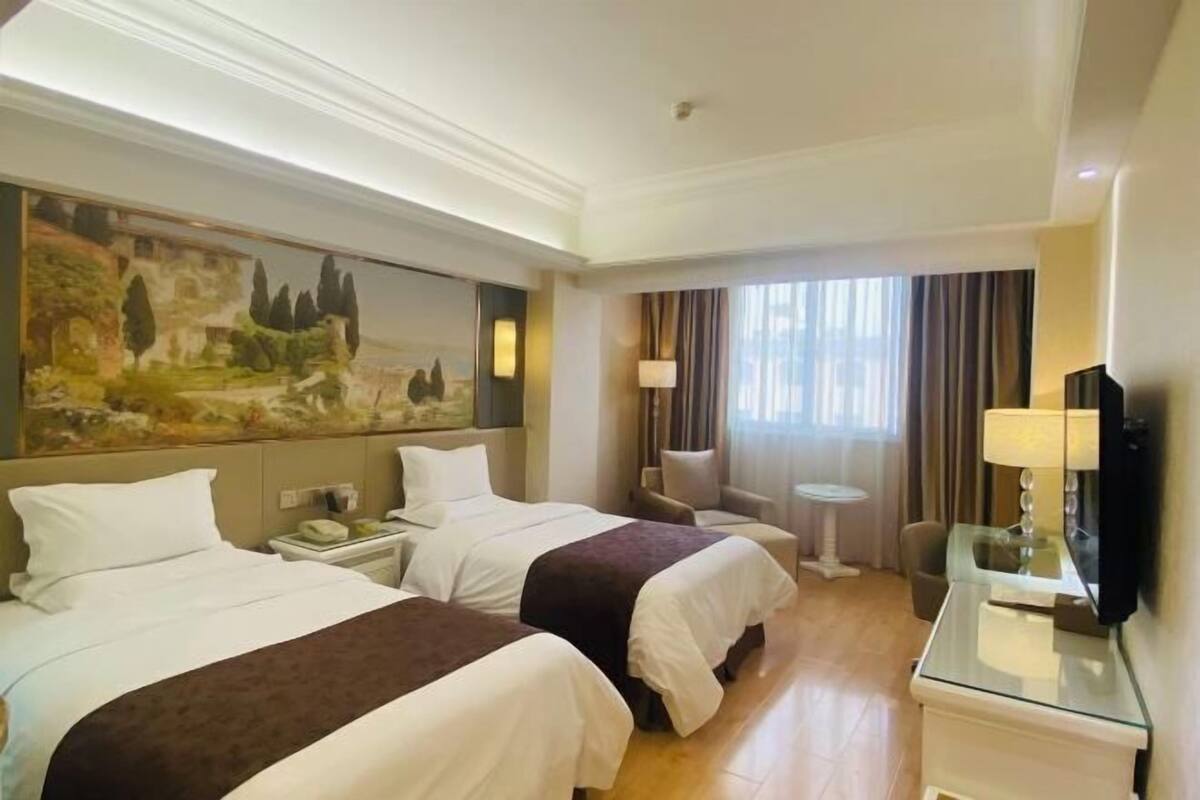 A hotel room with two beds, a painting on the wall, a television, and a small table with a lamp.
