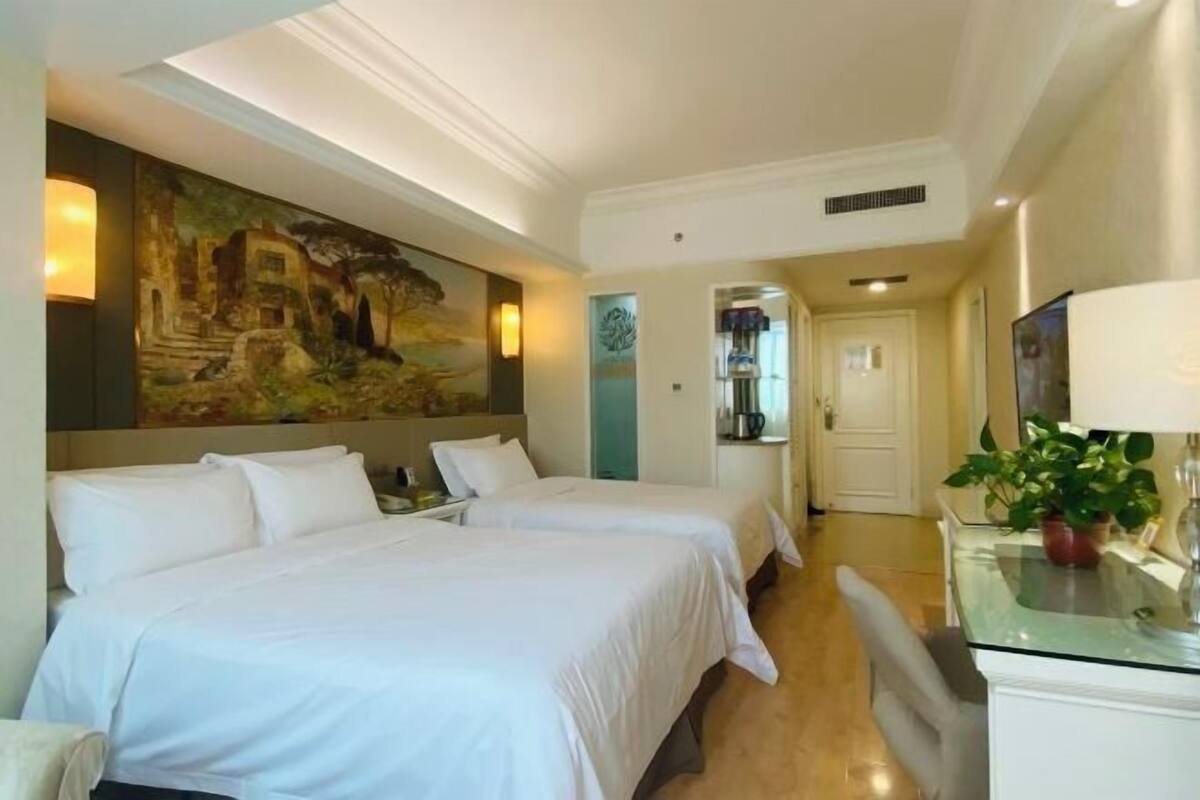 A hotel room with two beds, a painting on the wall, a desk with a plant, and a door leading to another room.