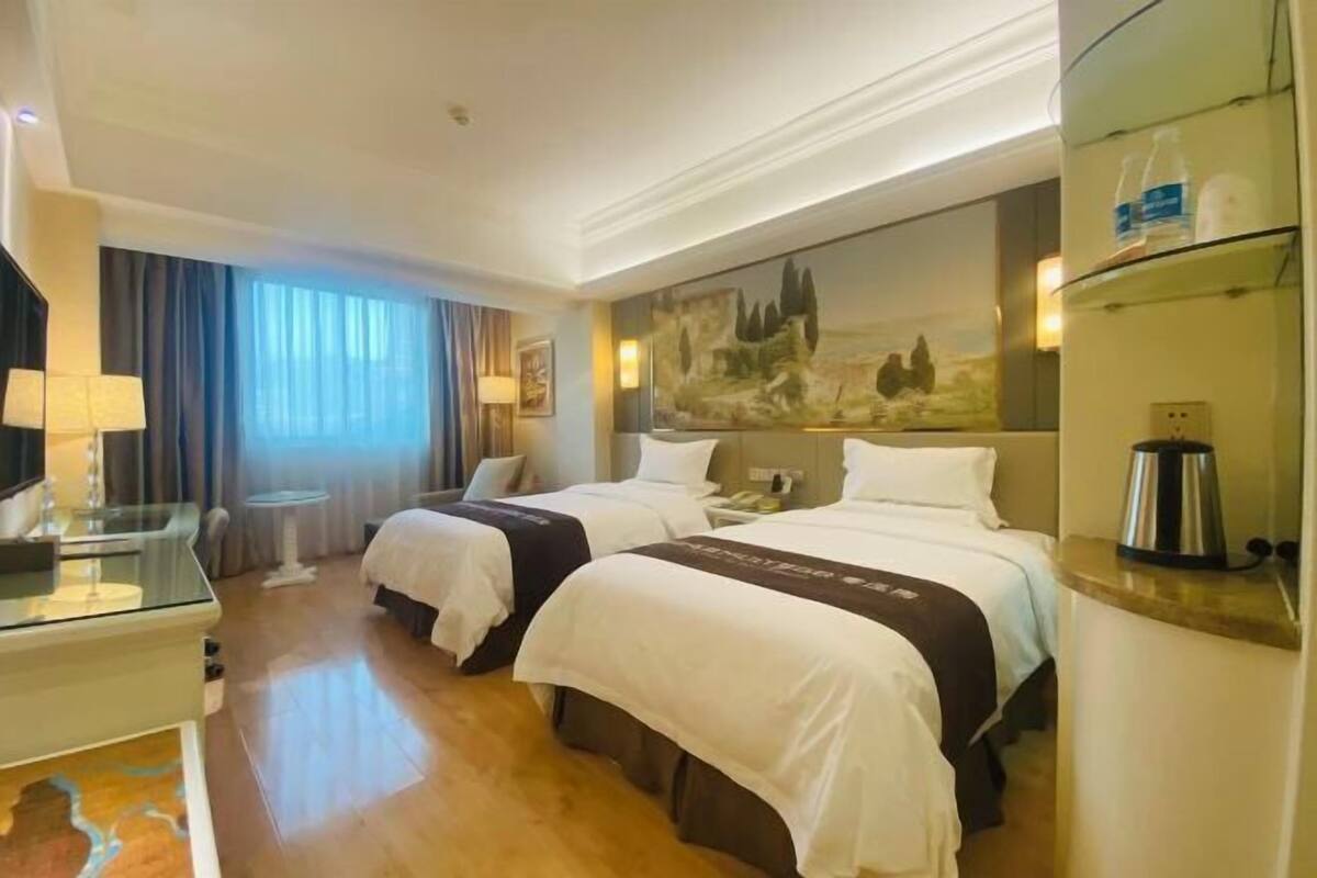 A hotel room with two beds, a desk, a chair, a painting on the wall, and a shelf with bottles.