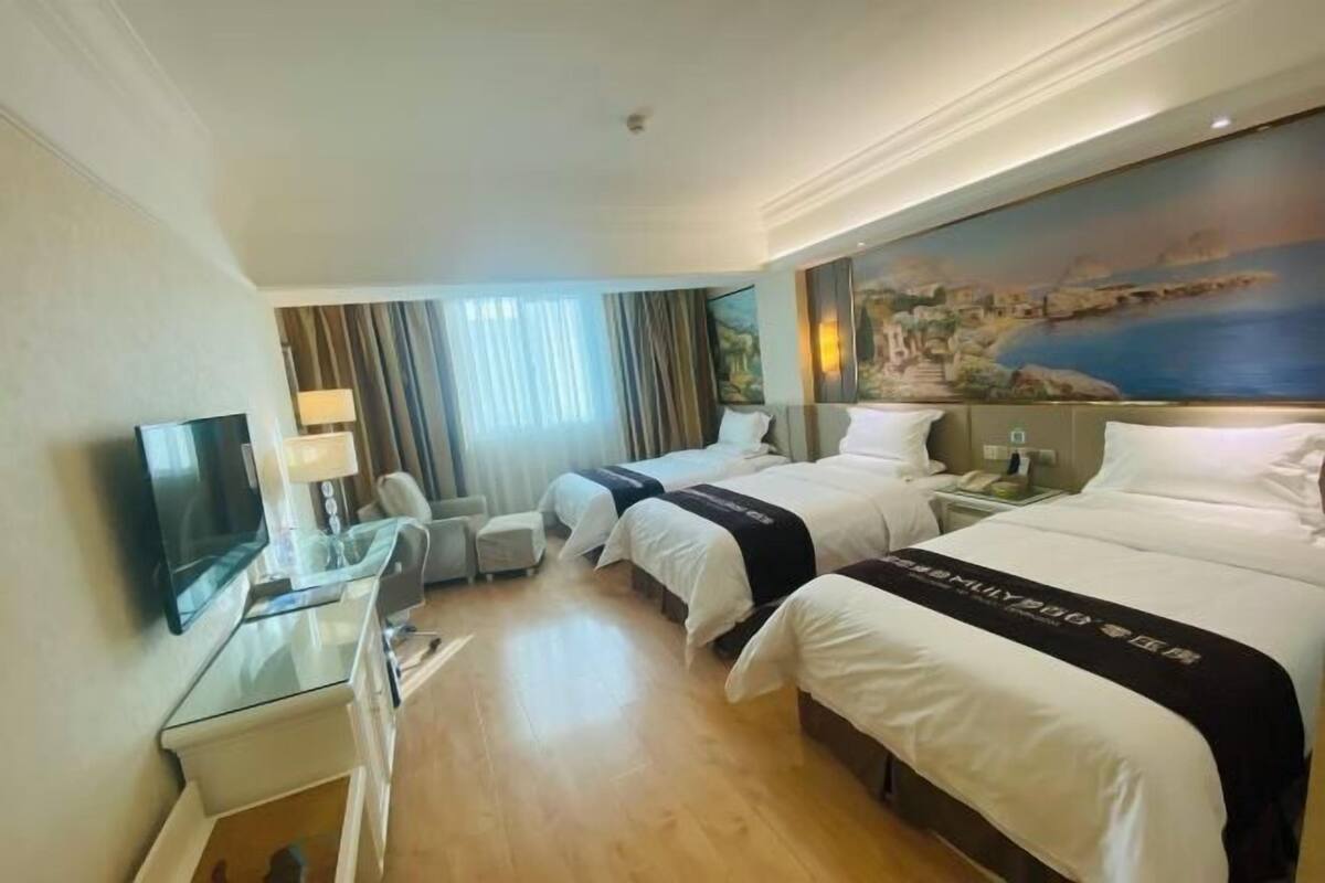 A hotel room with two beds, a TV, a desk, and a painting of a coastal scene.