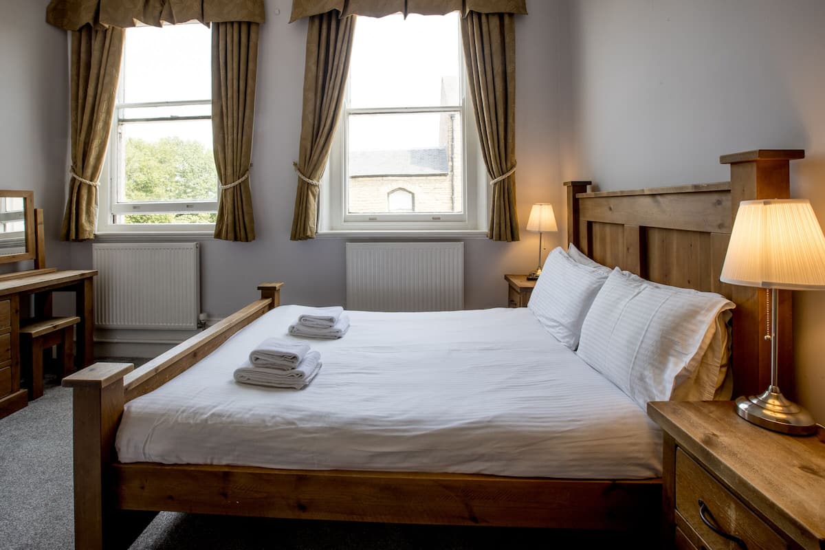 A neatly made bed with white linens and towels, a wooden headboard, a bedside lamp, and a view of the outdoors through a window with curtains.