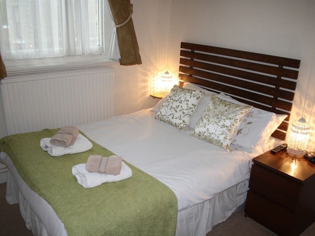 Una cama bien tendida con un edredón verde, dos toallas y almohadas estampadas. Hay lámparas de noche a ambos lados de la cama.