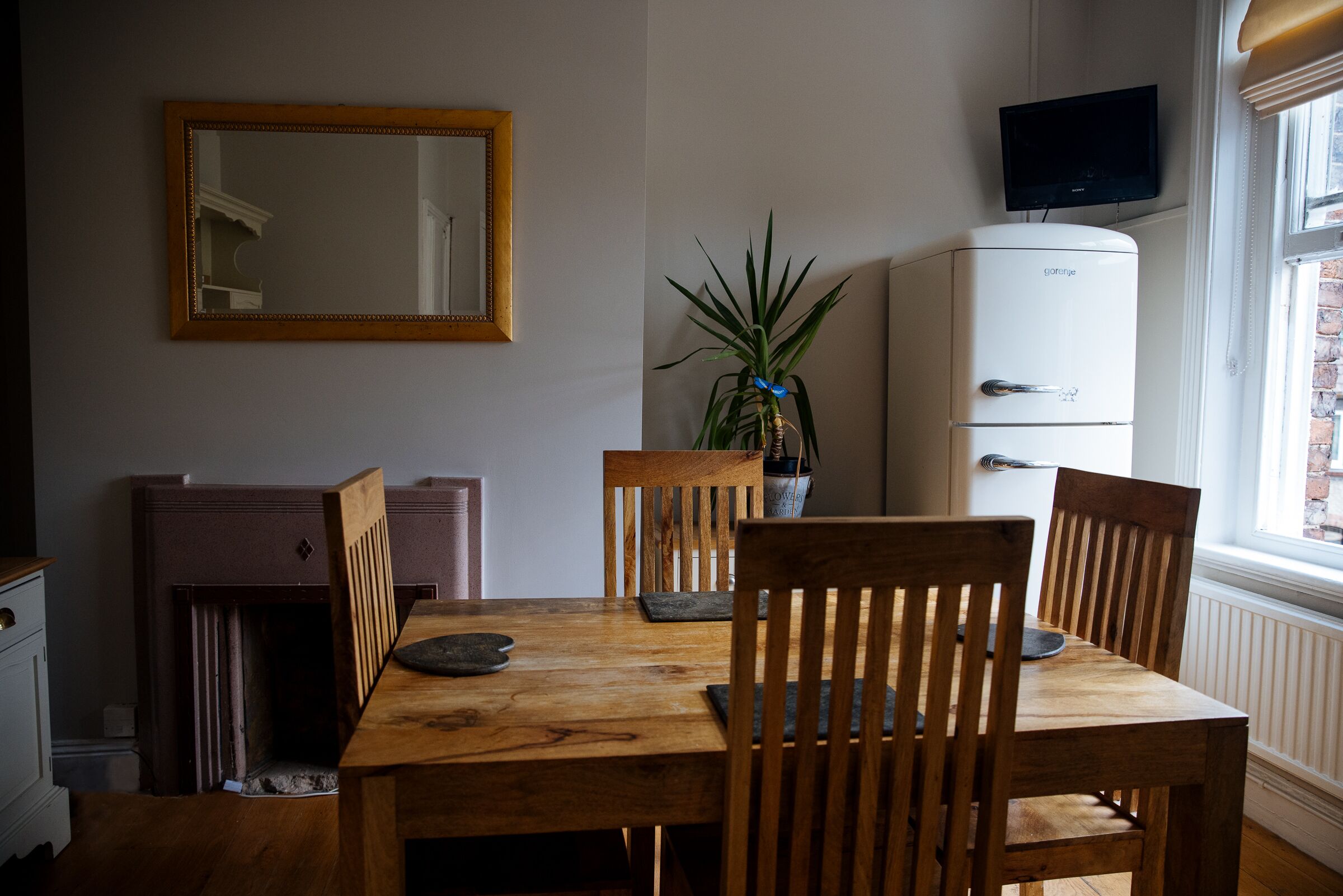 Un coin repas avec une table et des chaises en bois, une plante en pot, un réfrigérateur blanc et un téléviseur fixé au mur.