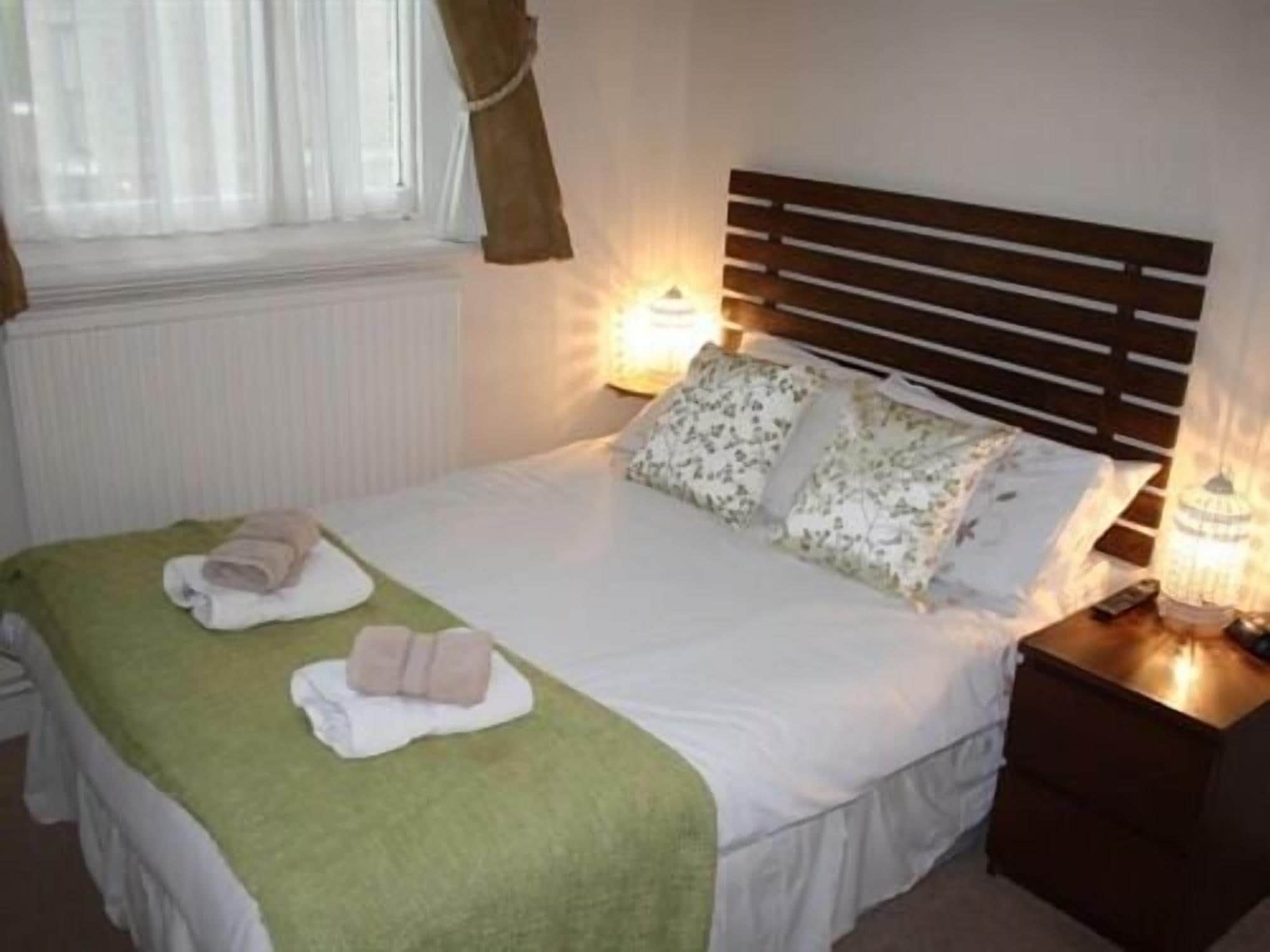 A neatly made bed with a green blanket and two folded towels, a wooden headboard, a bedside lamp, and a window with curtains.