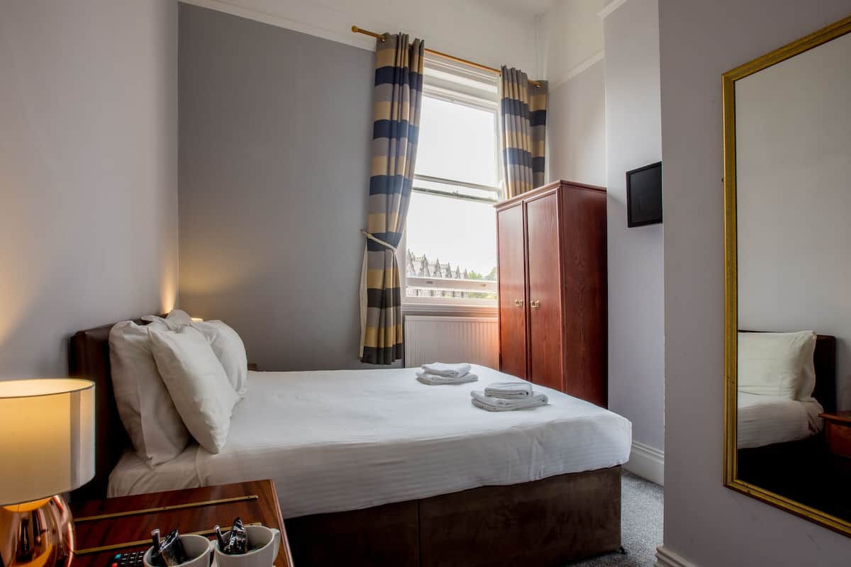 A hotel room with a bed, a wooden wardrobe, a mirror, and a window with curtains.