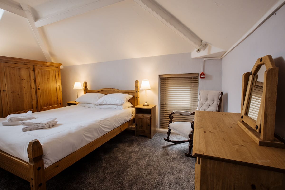 A bedroom with a wooden bed, two bedside lamps, a rocking chair, a wooden dresser, and a window with blinds.