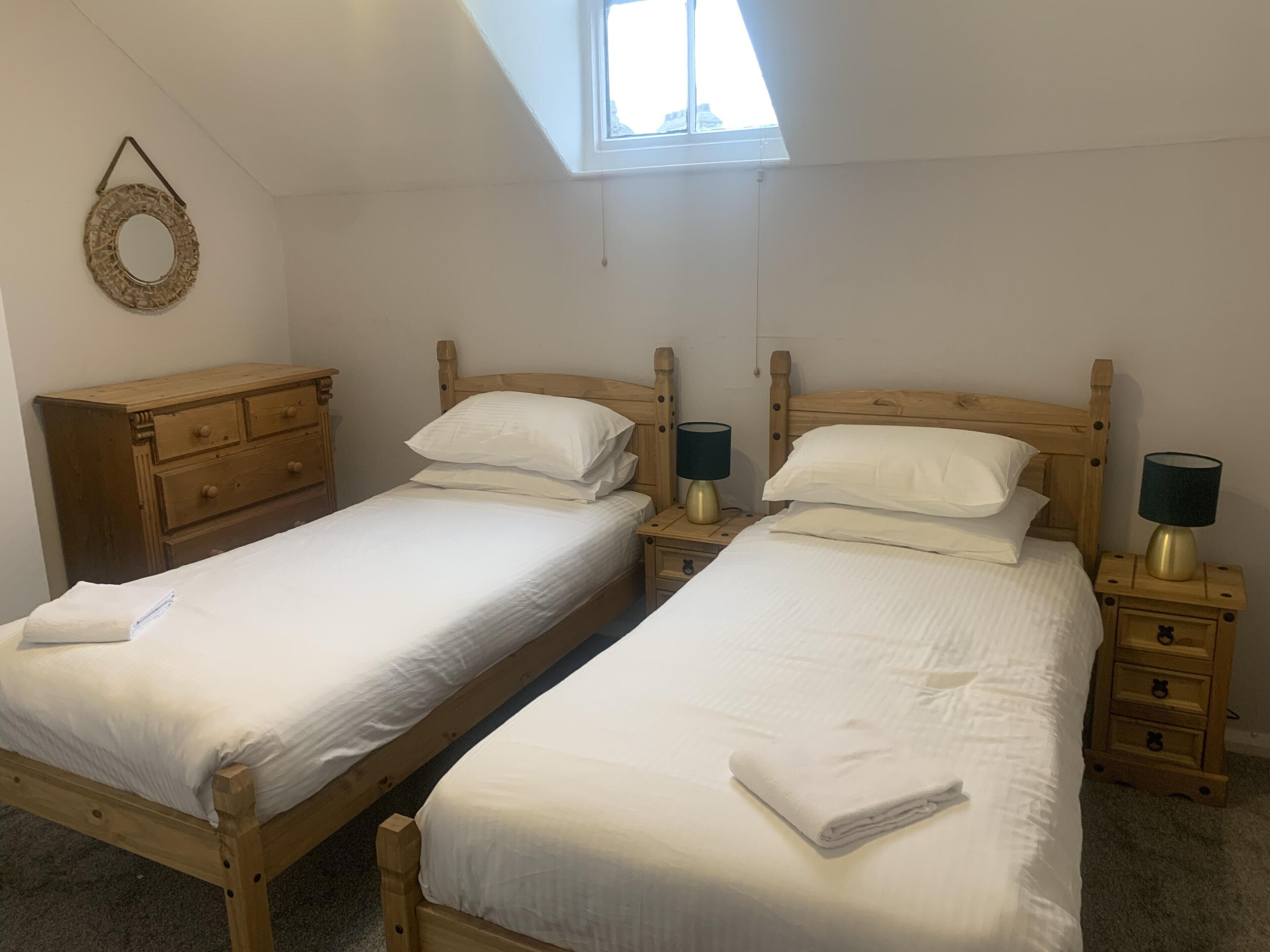 Une chambre à coucher avec deux lits en bois, chacun muni d’une table de chevet et d’une lampe, une commode en bois et une lucarne au plafond.