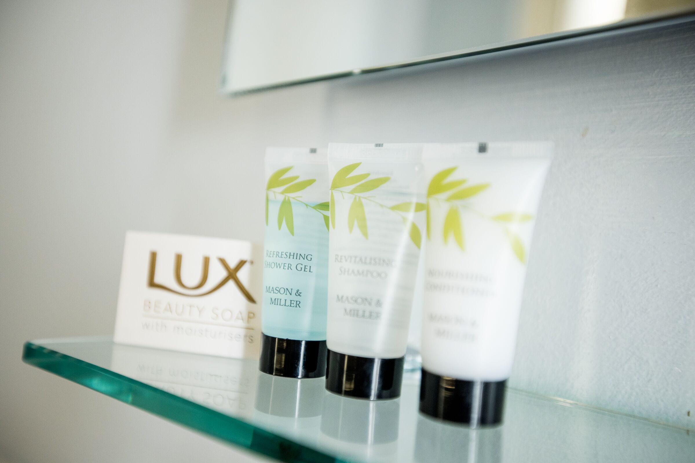 A glass shelf with three bottles of Lux Beauty Soap and two bottles of Mason & Miller shower gel.