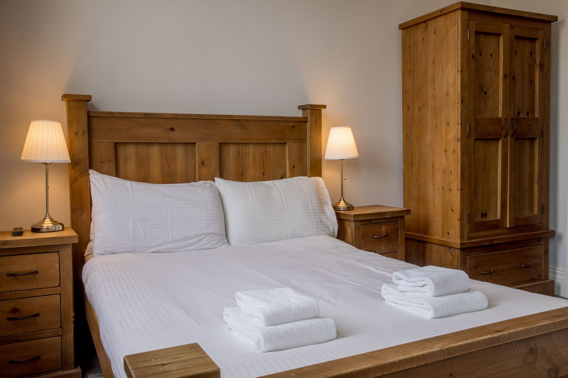 A neatly made bed with white linens and two bedside lamps. There is a wooden wardrobe to the right of the bed.
