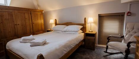 A bedroom with a wooden bed, two bedside tables with lamps, a rocking chair, and a window with blinds.