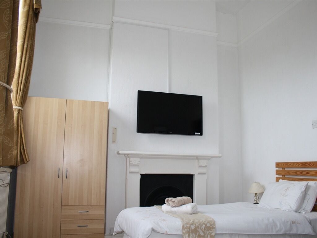 A bedroom with a bed, a television mounted above a fireplace, wooden wardrobe, and a nightstand with a lamp.