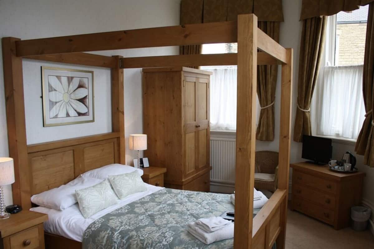 A four-poster bed with a floral bedspread, a bedside table with a lamp, a wooden wardrobe, and a television on a wooden dresser.