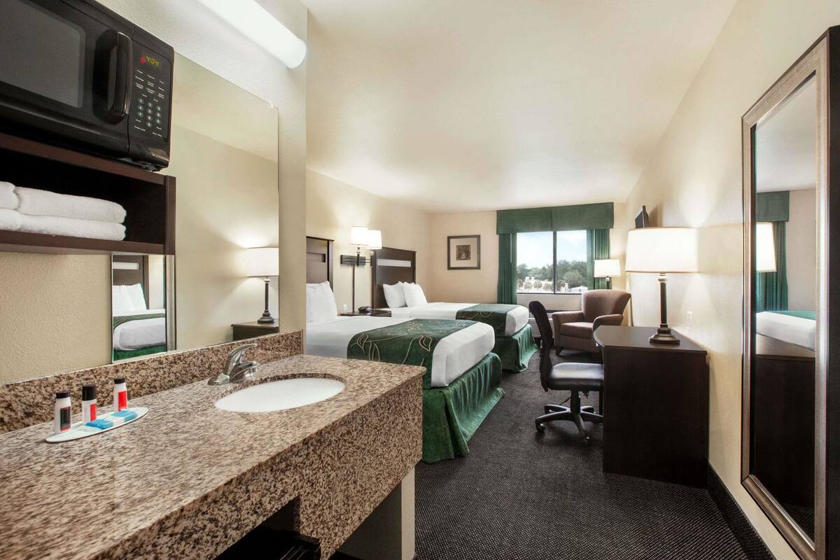 A hotel room with a granite countertop sink, two beds with green bedding, a desk with a chair, and a large mirror.