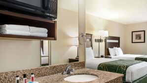 Quarto de hotel com lavatório de bancada de granito, um espelho, televisão sobre o balcão, cama com roupa de cama verde, mesa de cabeceira com luminária e um quadro na parede.